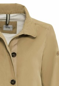 Béžová bunda s jemnou texturou, s límcem, dvěma tmavými knoflíky a nášivkou s logem na levém rukávu.