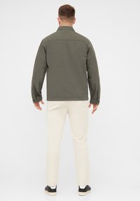 Chaqueta de camisa verde oscura con cuello, mangas largas y puños abotonados, combinada con pantalones color crema y zapatillas negras.