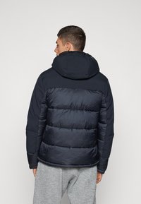 Marineblå pufferjakke med hætte, der har quiltede sektioner og en glat overflade. Båret over grå sweatpants, hvilket viser en afslappet pasform.