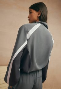 Gray athletic jacket with white side stripes, relaxed fit, long sleeves, and a high collar. Smooth texture with minimalistic design.