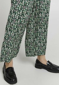 Blommönstrade vida byxor i mörkgrön, med små vita och rosa blommor. Svarta blanka loafers med tjock sula kompletterar looken.