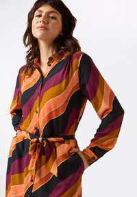 Kleid mit Knopfleiste, das auffällige, wellenförmige Streifen in Orange, Lila und Schwarz zeigt. Lange Ärmel und ein Bindegürtel betonen die Taille. Glatte Stofftextur.