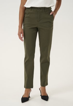 Personne portant un pantalon droit couleur olive et des talons noirs à bout pointu, debout avec une main dans la poche devant un fond uni.