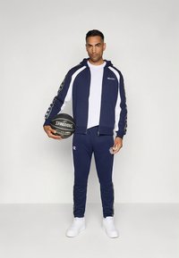 Tamsiai mėlynas sportinis kostiumas su baltais akcentais, sudarytas iš užtraukiamos striukės ir atitinkančių kelnių. Laiko krepšinio kamuolį, ant aprangos matomi logotipo elementai.