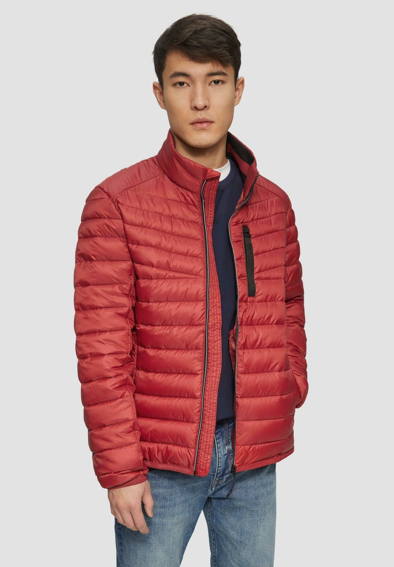 Giovane uomo che indossa una giacca trapuntata rossa sopra un felpa blu navy e jeans blu, in piedi con una mano in tasca davanti a uno sfondo neutro.