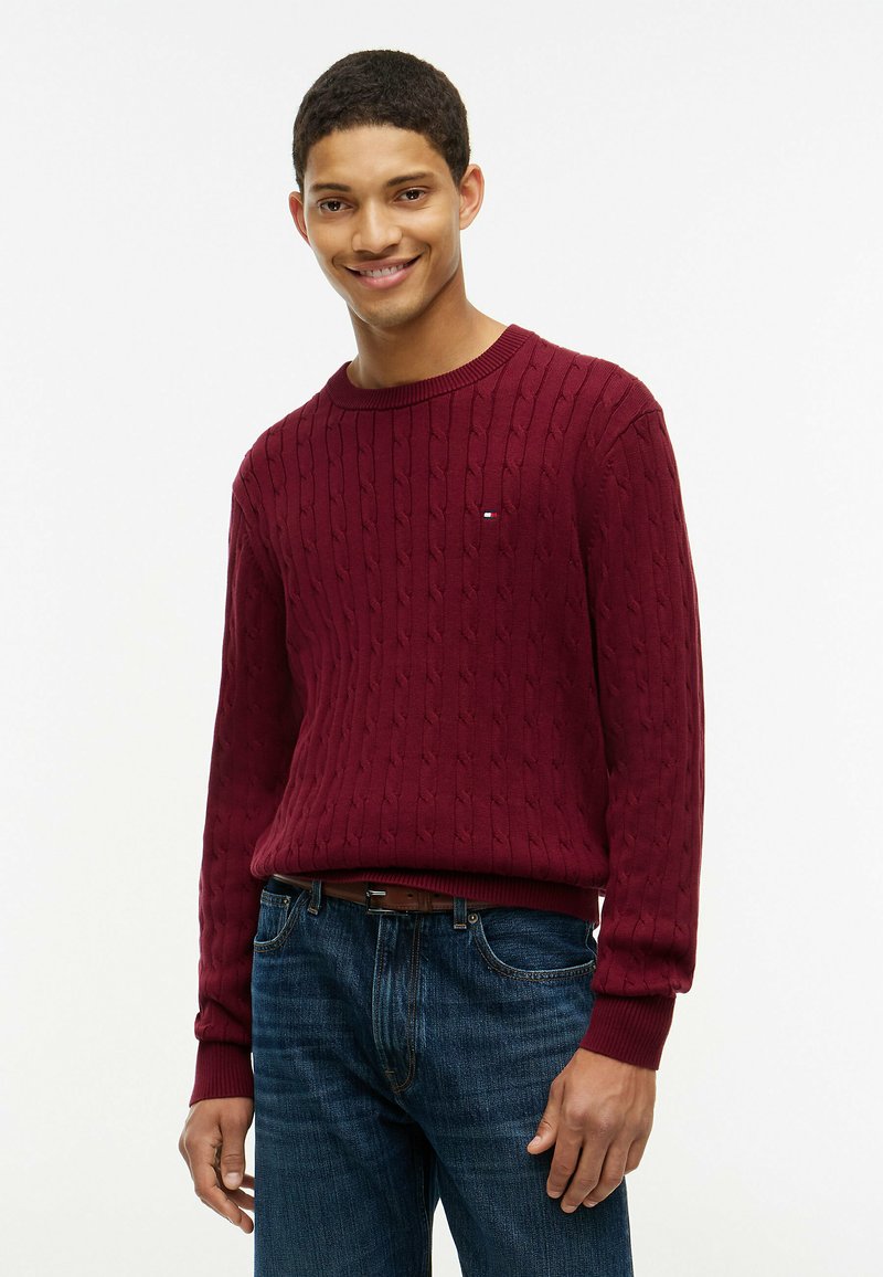 Jeune homme souriant, portant un pull torsadé rouge et un jean bleu, debout devant un fond blanc uni.