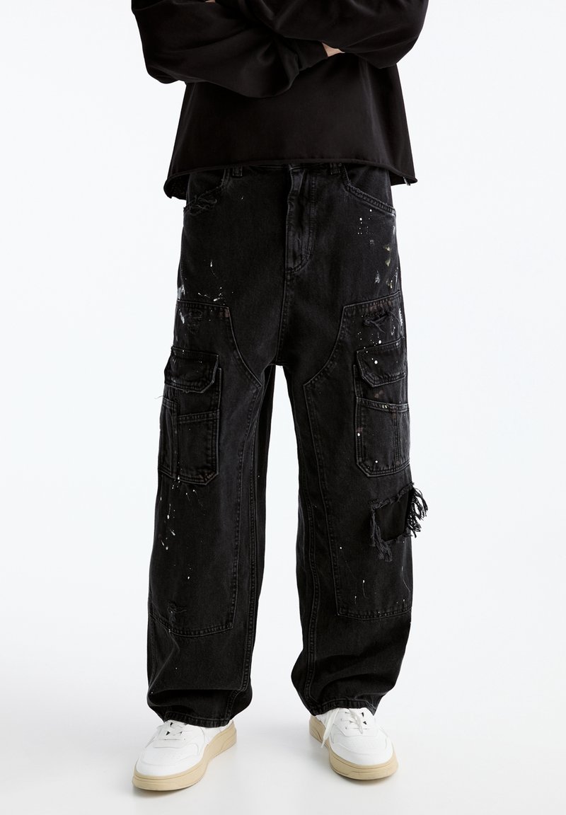 Pantalones cargo anchos negros con detalles desgastados y manchas de pintura, combinados con zapatillas blancas y una camiseta de manga larga negra.