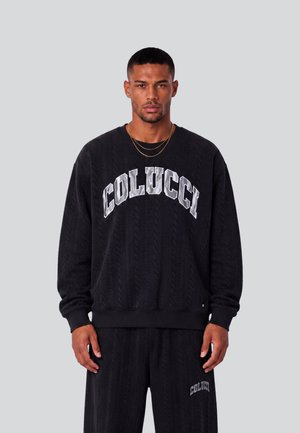 Mann trägt schwarzen Colucci-Sweatshirt und passende Hose mit Zopfmuster, steht vor schlichtem grauem Hintergrund.