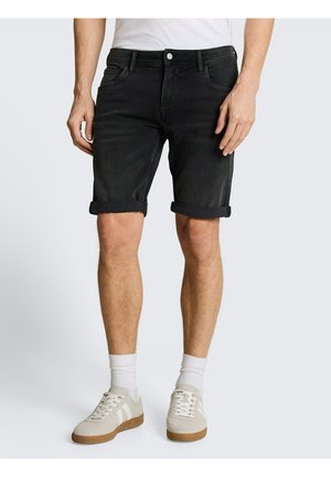Man draagt opgerolde zwarte denim shorts, witte sokken en beige sneakers, staand tegen een effen achtergrond.