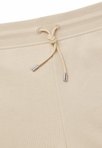 Beige Jogginghose mit einem Kordelzugbund, ausgestattet mit silberfarbenen Aglets. Glatte Textur mit elastischem Abschluss für einen engen Sitz.