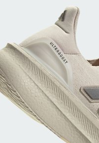 Primer plano de unas zapatillas de running Adidas Ultraboost en tono beige, mostrando la suela texturizada, la parte superior de tela, el soporte en el talón y el texto del logo en el contrafuerte del talón.