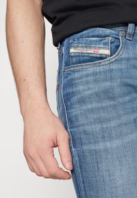 Jean en denim bleu avec une coupe fuselée, délavage clair, coutures contrastées et un patch logo sur la poche arrière.