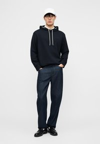 Νέος άνδρας που φοράει λευκό καπέλο, μαύρο φούτερ με κορδόνια, σκούρο μπλε τζιν και μαύρα παπούτσια, στέκεται με τα χέρια στις τσέπες μπροστά σε απλό φόντο.