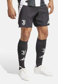 Pantaloni sportivi neri con dettagli bianchi e logo della Juventus, abbinati a calze lunghe nere con il logo e scarpe da ginnastica bianche.