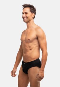 Schwarze Baumwollslips mit einem enganliegenden Design, ausgestattet mit einem breiten Bund und ohne sichtbare Muster. Das Model steht mit einem neutralen Gesichtsausdruck.