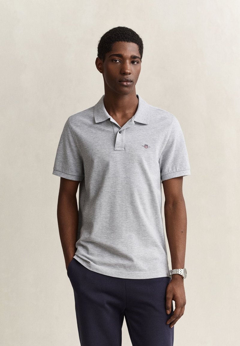 Jeune homme portant un polo gris clair et un pantalon foncé, debout avec une main dans la poche sur un fond neutre.