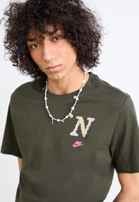Oliven grøn bomulds t-shirt med et tekstureret 'N' logo i beige og et rosa Nike swoosh. Båret med en perle halskæde.