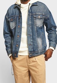 Giacca di jeans di colore azzurro chiaro con una finitura sbiadita, due tasche sul petto, bottoni color rame e una vestibilità comoda, indossata sopra un hoodie bianco.