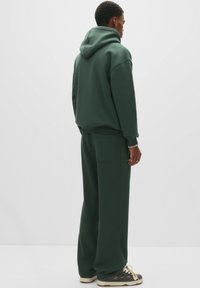 Mann in einem dunkelgrünen Hoodie und passenden weit geschnittenen Hosen, mit grauen und weißen Sneakers, steht mit dem Rücken zu einem weißen Hintergrund.