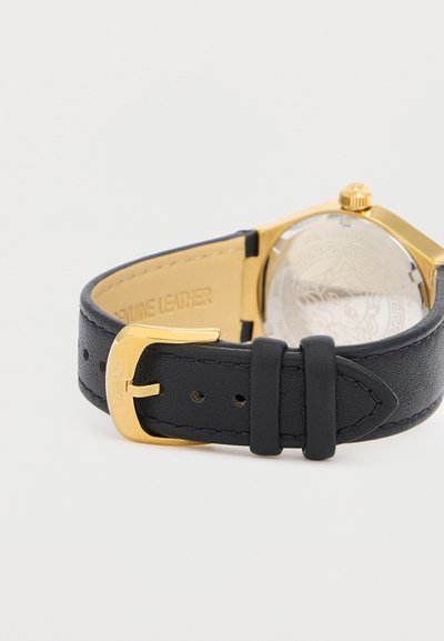 Bracelet de montre en cuir noir véritable avec boucle dorée et boîtier de montre doré, fond gris clair uni.