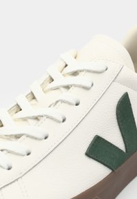White leather sneaker featuring a green suede logo, flat white laces, and a brown rubber sole. Close-up of the upper design and texture.