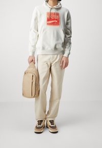 Sudadera blanca con gráfico rojo, pantalones beige y zapatillas con detalles en marrón. Sosteniendo un bolso cruzado color beige con compartimentos con cremallera.