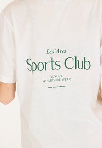 T-shirt en coton blanc avec un texte vert qui dit "Les'Arcs Sports Club Luxury Athleisure Wear" et "Nouvelles Mauvaises Habitudes" en dessous.
