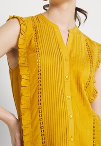 Hnědožlutá bezrukávová blůza s vertikálními sklady, volánkovými rukávy a dekorativním krajkovým lemováním kolem knoflíků. Hladká textura.