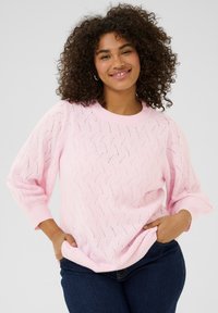 Pull en tricot rose clair avec un motif de zigzags et de trous, col rond et manches amples. Associé à un jean en denim foncé.