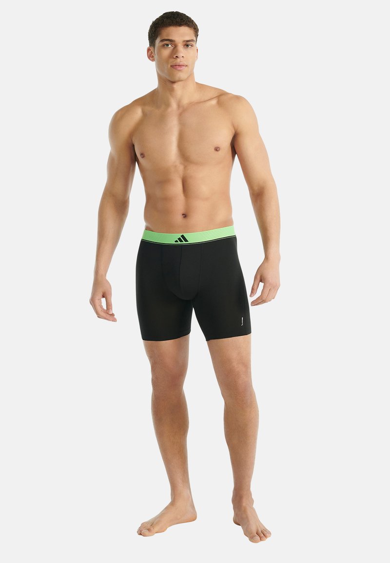 Svalnatý muž stojící bos v černých boxerkách se zeleným páskem a logem Adidas na přední straně, na jednobarevném pozadí.