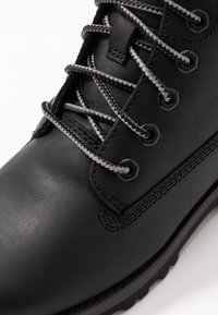 Bota de cuero negro con ojales metálicos, cordones grises y costuras texturizadas. La suela es resistente para un mejor agarre.