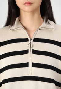 Suéter beige con rayas horizontales negras, cuello con cremallera y tirador de cremallera plateado. Presenta una textura acanalada y un corte relajado.