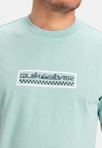 Světle tyrkysové tričko s krátkým rukávem vyrobené z bavlny, se silným černobílým designem loga a kostkovanými akcenty na přední straně.