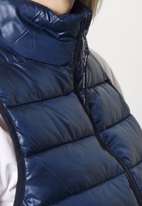 Marineblaue Steppweste mit hohem Kragen, Reißverschluss und gesteppter Textur. Verfügt über eine glatte, glänzende Oberfläche und kontrastierender schwarzer Verzierung.