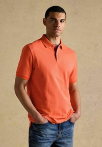 Jeune homme debout, mains dans les poches, portant un polo à manches courtes orange et un jean bleu, sur un fond beige uni.