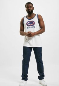 Witte tanktop met blauwe rand en grafisch logo; gecombineerd met donkere spijkerbroek en witte sneakers, wat een casual outfit toont.