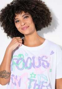 Hvid kortærmet t-shirt med farverig graffiti-lignende tekst og illustrationer, herunder hjerter, stjerner og en diamant, på et glat stof.