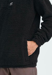 Schwarzer Fleece-Pullover mit einer strukturierten, weichen Oberfläche. Mit einer Fronttasche und gerafften Ärmelbündchen. Etikett im Brustbereich.