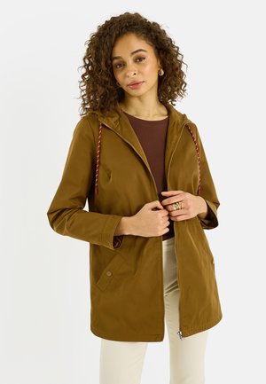 Mujer con cabello rizado que lleva una chaqueta marrón con cordones rojos, una blusa marrón oscuro, pantalones crema y joyería dorada, sobre un fondo liso.