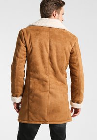 Chaqueta de ante color tostado con un suave cuello y puños de borrego blanco, corte recto, con costuras verticales y una textura suave.