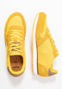 Gelbe Sportschuhe aus Mesh- und Wildledermaterialien, mit runder Zehenpartie, gepolstertem Kragen, Korksohle und weißer Gummisohle mit strukturierten Details.