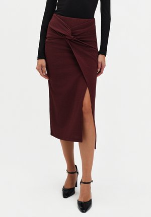 Bordowa spódnica do połowy łydki z marszczonym przodem i rozcięciem po boku, wykonana z teksturowanej tkaniny; w połączeniu z czarnymi butami na wysokim obcasie.