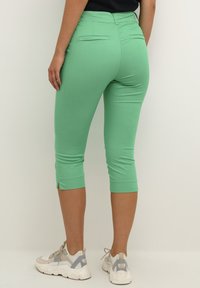 Green cropped leggings with a smooth texture, featuring two back pockets and side slits at the hem, paired with light-colored sneakers.