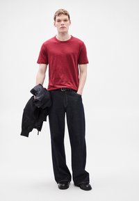 Κόκκινο κοντομάνικο μπλουζάκι, σκούρο τζιν παντελόνι με φαρδιά πόδια, μαύρο μπουφάν bomber κρατημένο στο χέρι, με λεπτή μαύρη ζώνη και μαύρα παπούτσια.