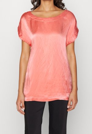 Vrouw die een losse, koraalroze satijnen blouse met korte mouwen en zwarte broek draagt, staand tegen een effen lichte achtergrond.