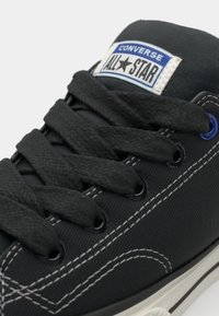 Black canvas sneaker with white stitching, textured laces, and a blue label reading "CONVERSE ALL-STAR." Circular black eyelets.