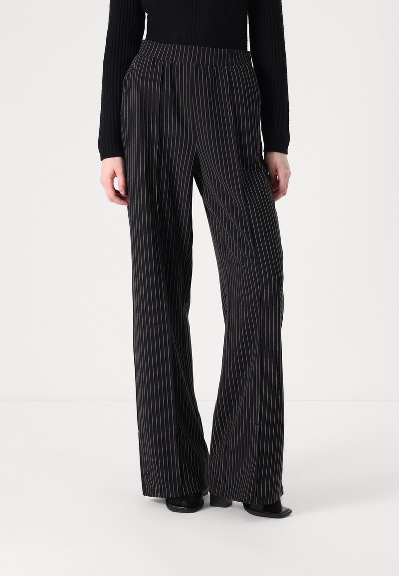 Femme portant un pantalon noir à jambe large avec de fines rayures blanches verticales et des chaussures noires, debout devant un fond clair uni.