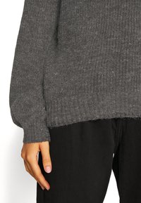 Maglione grigio a maglia con polsini e orlo a coste. Tessuto morbido e strutturato. Abbinato a pantaloni neri. Vista parziale di una mano che tiene l'orlo.