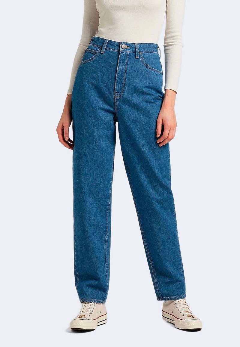 Personne portant un jean bleu taille haute, un haut crème à manches longues et des baskets blanches, debout devant un fond uni.