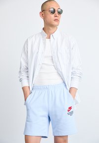Lysblå bomulds shorts med logo, hvid tanktop og lang, stribet skjorte med knapper. Runde solbriller fuldender outfittet.
